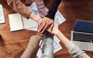 Group of colleagues overlaying hands with papers and laptop on desk beneath