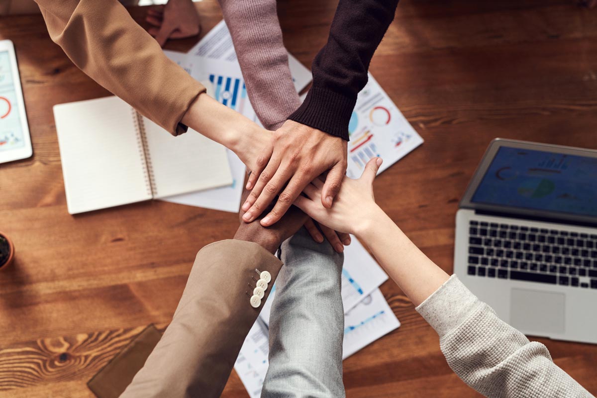 Group of colleagues overlaying hands with papers and laptop on desk beneath