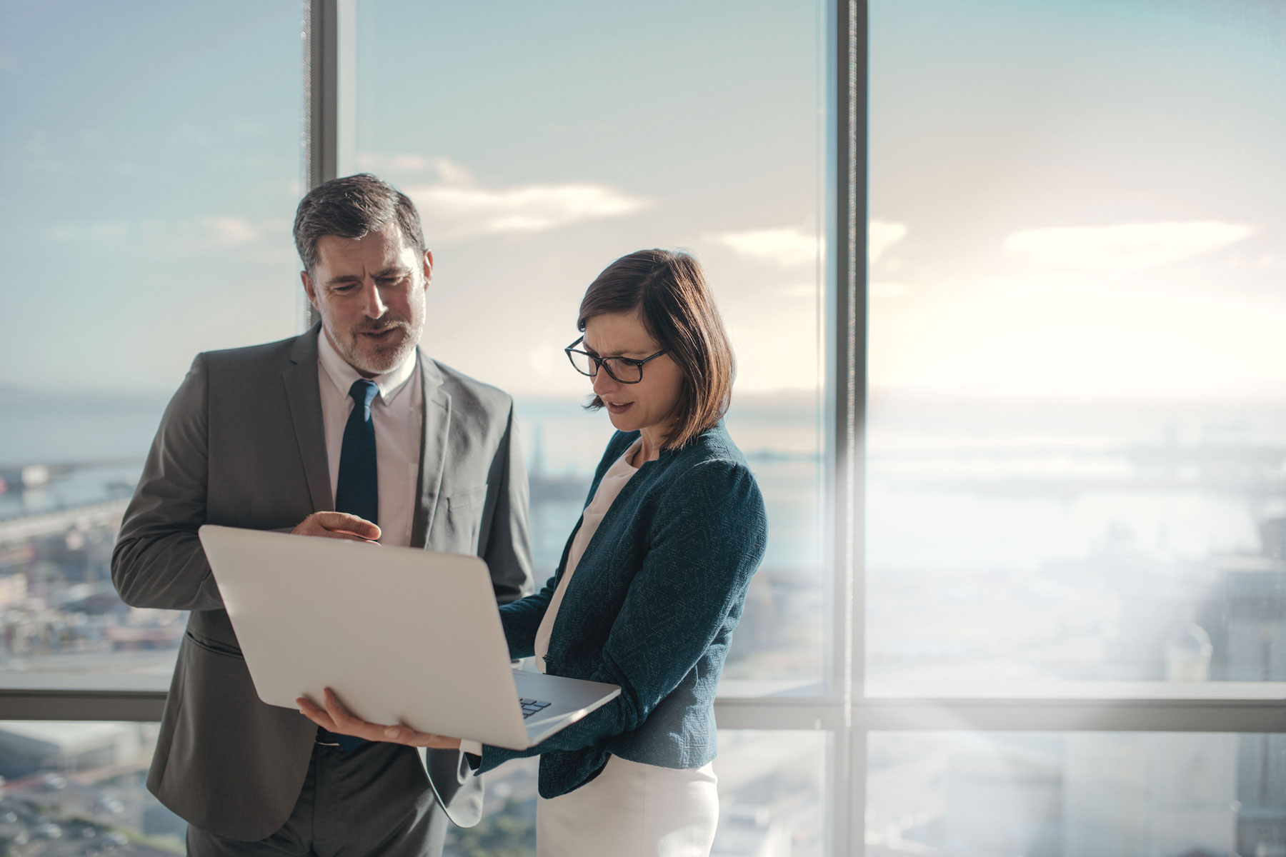 male and female collegue standing in front of window looking at laptop