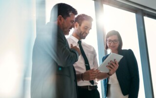 Three colleagues standing near a window looking at a laptop