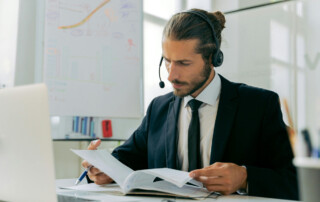 Business person looking at paperwork while seated behind a laptop with a headset on.