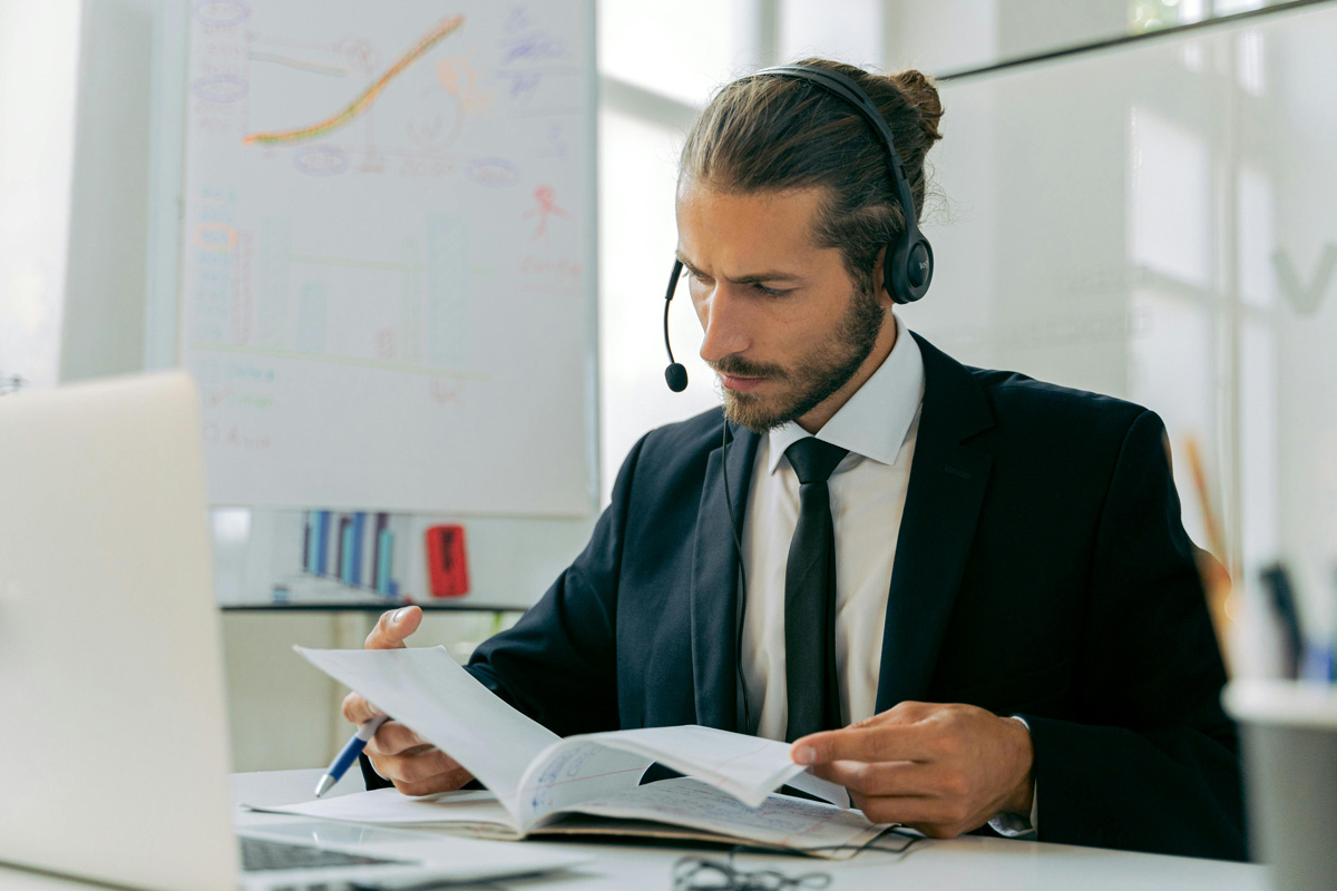 Business person looking at paperwork while seated behind a laptop with a headset on.