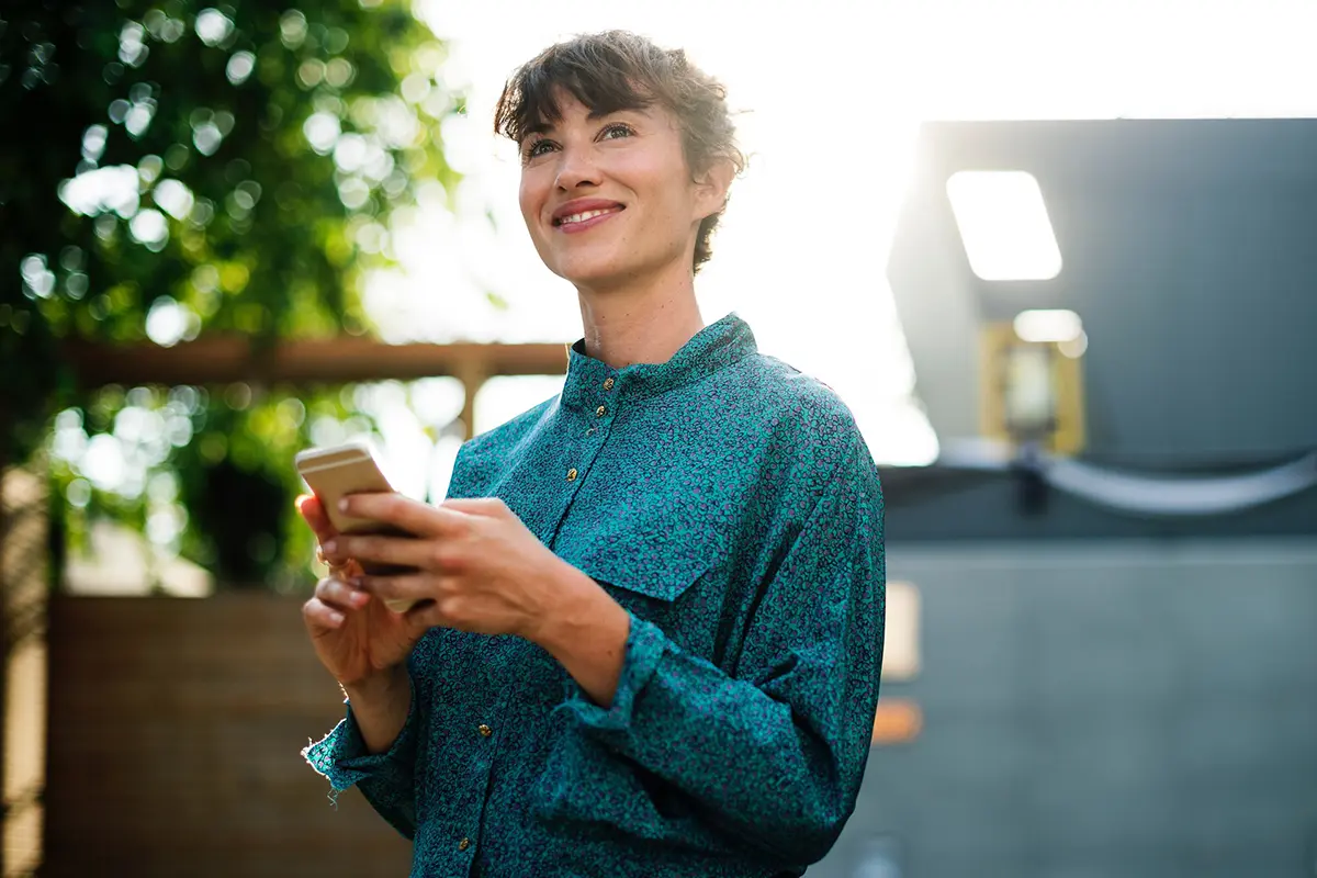 Person in teal button-up shirt using a smartphone outdoors, with bright sunlight and greenery in the background — representing mobile connectivity and outdoor lifestyle