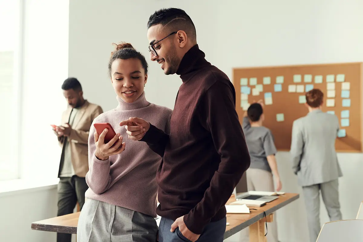 Group of people in an office: two in the foreground interacting with a red smartphone, and two in the background near a corkboard covered in sticky notes — highlighting teamwork, mobile technology, and collaborative planning.