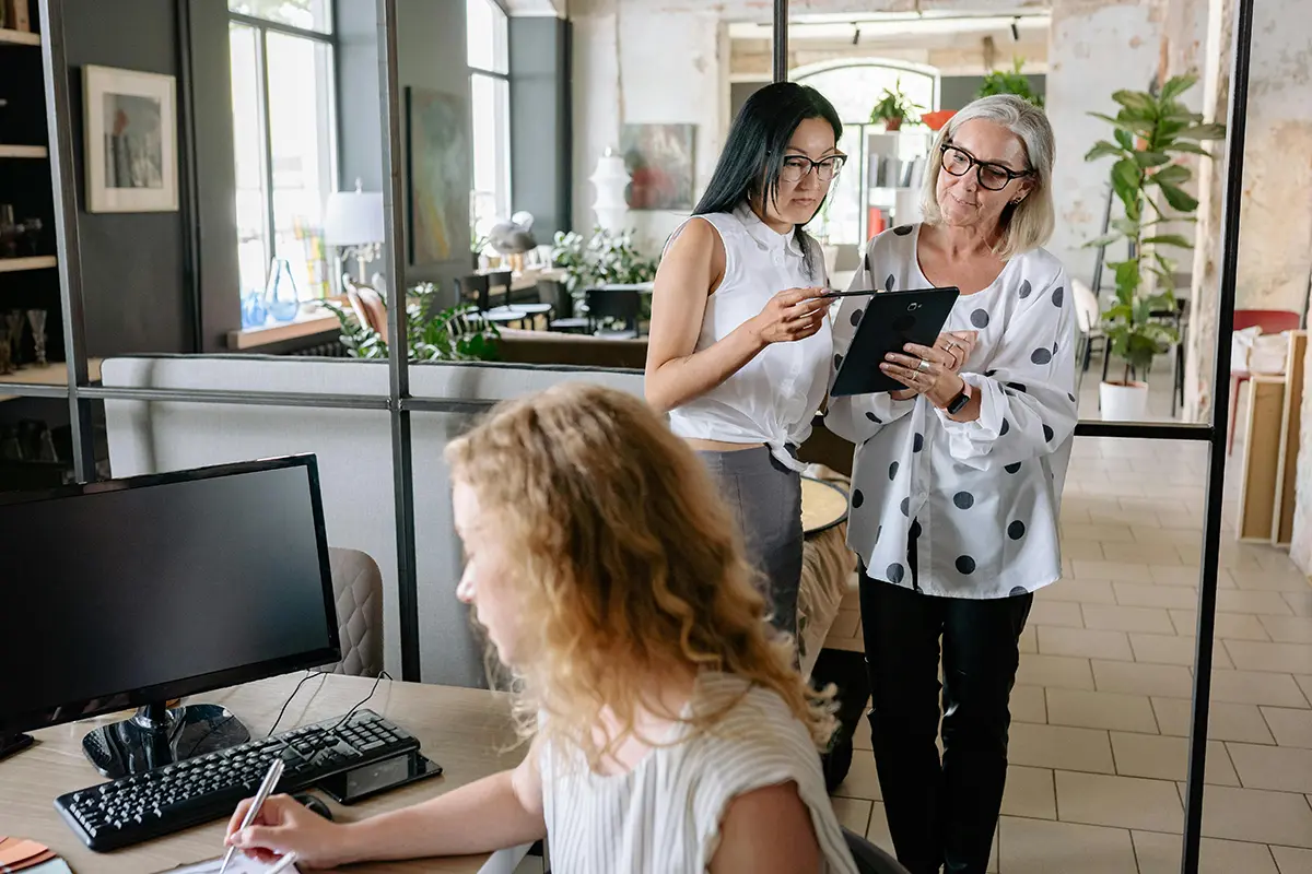 Three people in a modern office: one seated at a desk writing on a notepad beside a computer, and two standing and viewing a tablet — showcasing teamwork, digital collaboration, and workplace productivity.